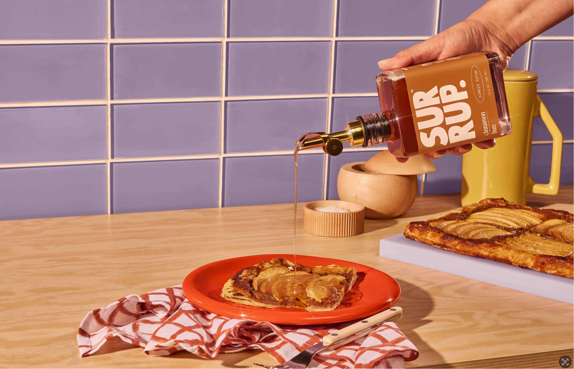 Person pouring syrup from a bottle labeled 'Sur Rup' onto pancakes on a plate with a blue tiled wall in the background.